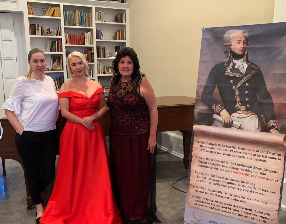 A photo of three women in concert attire in a musical salon next to a poster of Lafayette