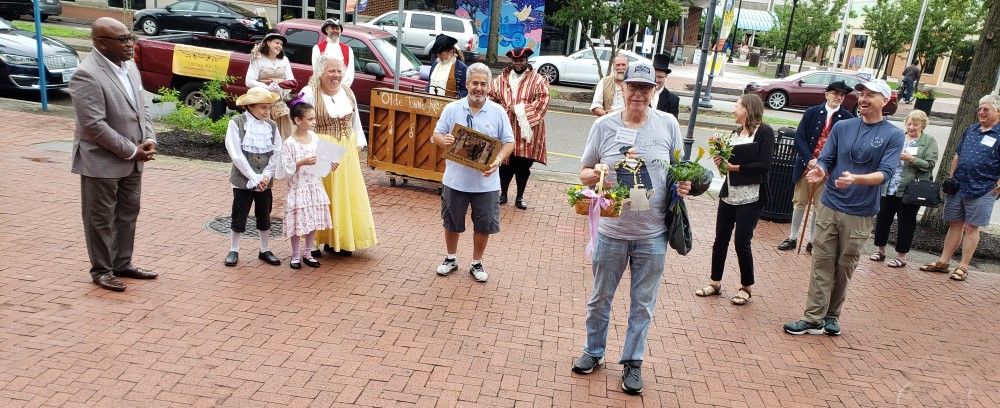 Portsmouth mayor (Mr.) Shannon Glover presents a plaque to AFL president Alan Hoffman; costumed historical interpreters, part of the troupe of Mary Veale and the Colonials, are in the background