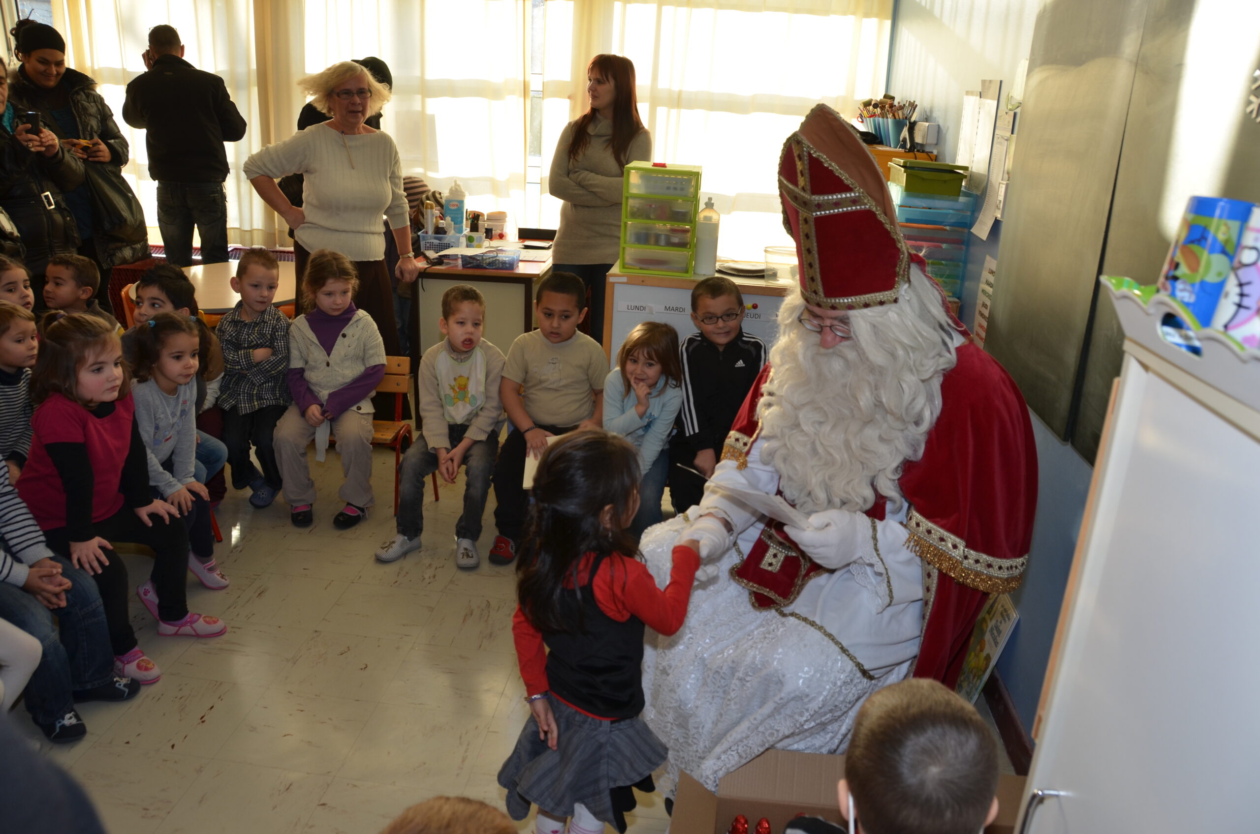 A photo of a group of schoolchildren in a classroom surrounding someone dressed as Saint Nicholas. In 2012 a group from Fayetteville visited a St Avold grade school on Dec 6, St Nicolas Day.